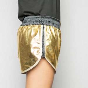 May include: Gold metallic shorts with a black waistband and white piping. The shorts are made of a shiny, reflective fabric.