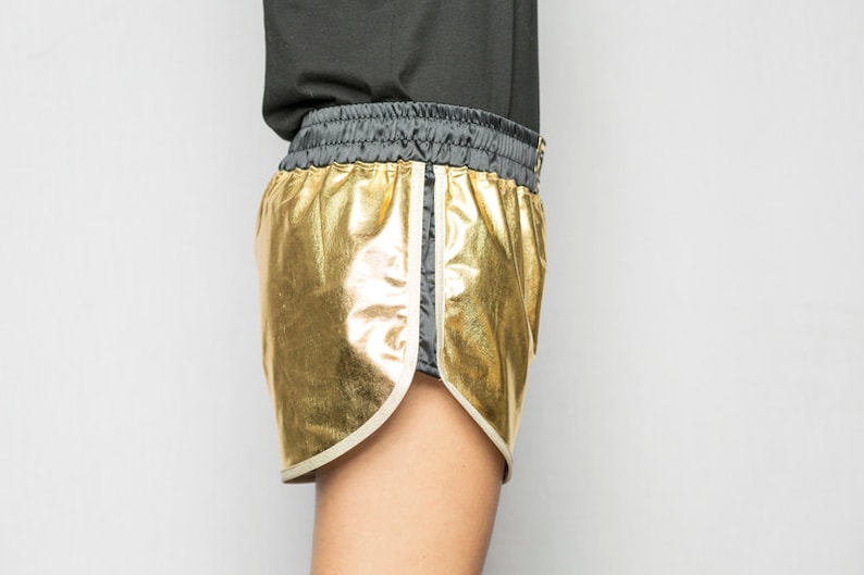 May include: Gold metallic shorts with a black waistband and white piping. The shorts are made of a shiny, reflective fabric.