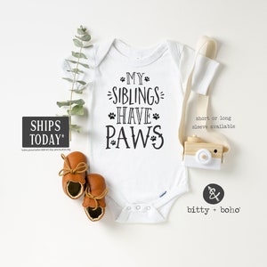 May include: White baby onesie with black text that reads "My siblings have paws" with paw prints. The onesie is on a white background with a wooden toy camera and brown baby shoes.
