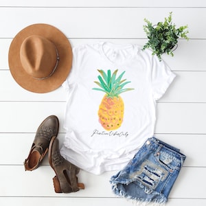 Op de afbeelding: Wit T-shirt met een aquarel ananas ontwerp en de tekst "Positive Vibes Only". Het shirt wordt gecombineerd met een spijkerbroekje, bruine laarzen en een bruine fedora.