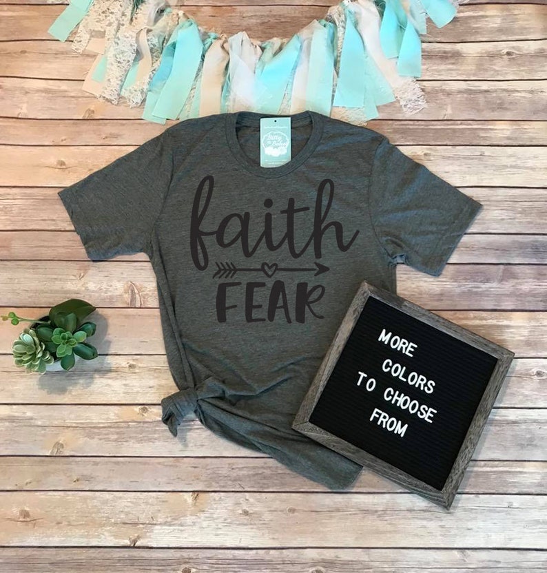 May include: A dark gray t-shirt with the words "faith" above "fear" in a black script font, with an arrow and heart design between the words. A framed letter board reads "MORE COLORS TO CHOOSE FROM". Decorative elements include a plant and a garland.