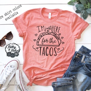 May include: A coral pink t-shirt with the text "I'm just here for the tacos" in black lettering with a black outline of a taco and hearts around it.