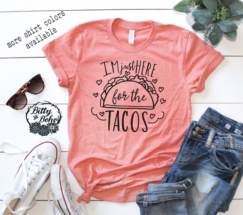 May include: A coral pink t-shirt with the text "I'm just here for the tacos" in black lettering with a black outline of a taco and hearts around it.
