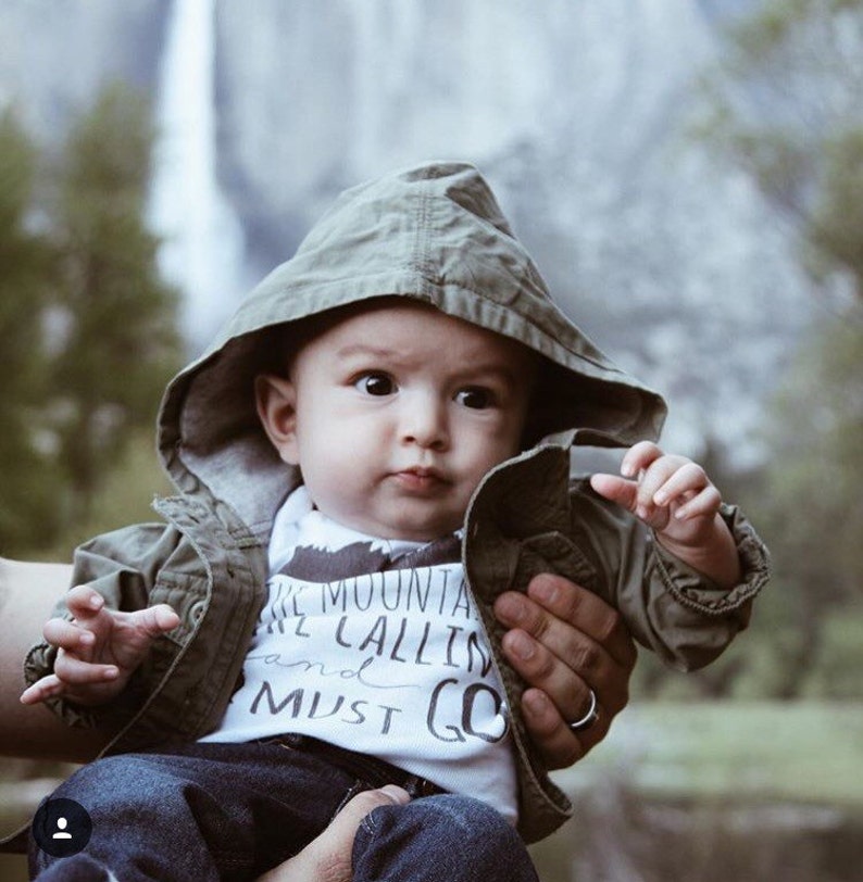 May include: A baby wearing a green hooded jacket and a white onesie with the text "The mountains are calling and I must go".