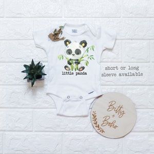 May include: White baby onesie with a panda bear graphic and the text "little panda". The onesie is available in short or long sleeve. The onesie is next to a wooden sign with the text "Bitty + Boho".