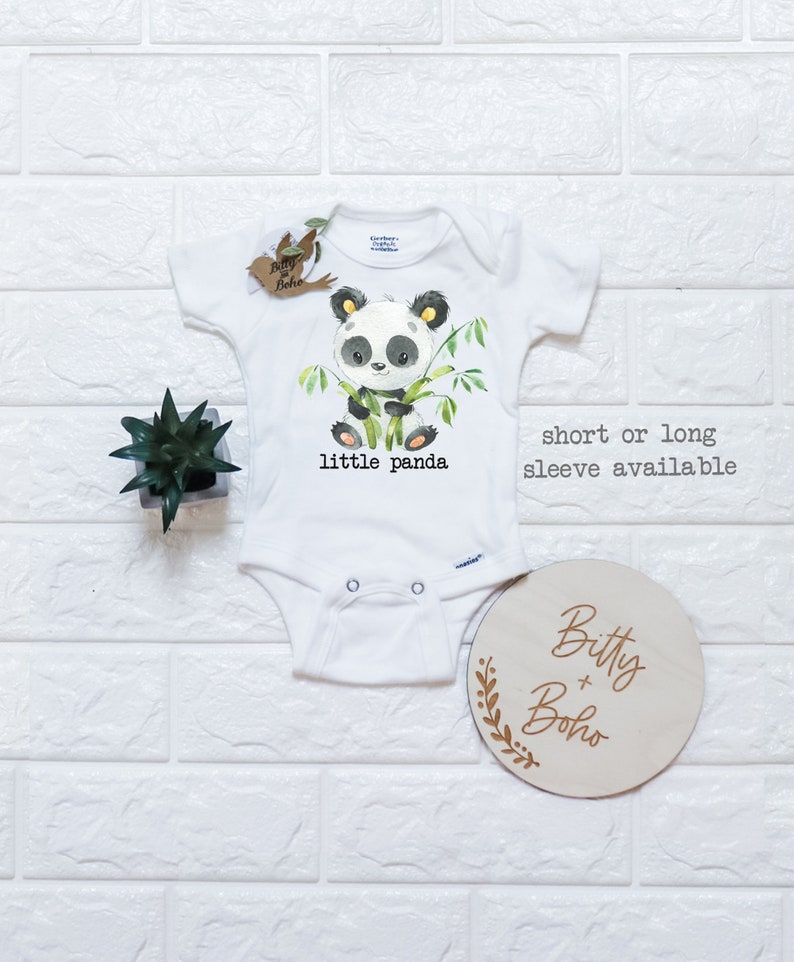 May include: White baby onesie with a panda bear graphic and the text "little panda". The onesie is available in short or long sleeve. The onesie is next to a wooden sign with the text "Bitty + Boho".
