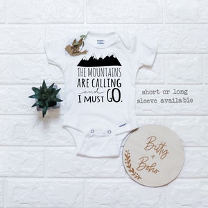 May include: White baby onesie with black text that reads "The Mountains Are Calling and I Must Go."  The onesie has a mountain graphic.  A small wooden sign with the text "Bitty + Boho" is in the lower right corner of the image.