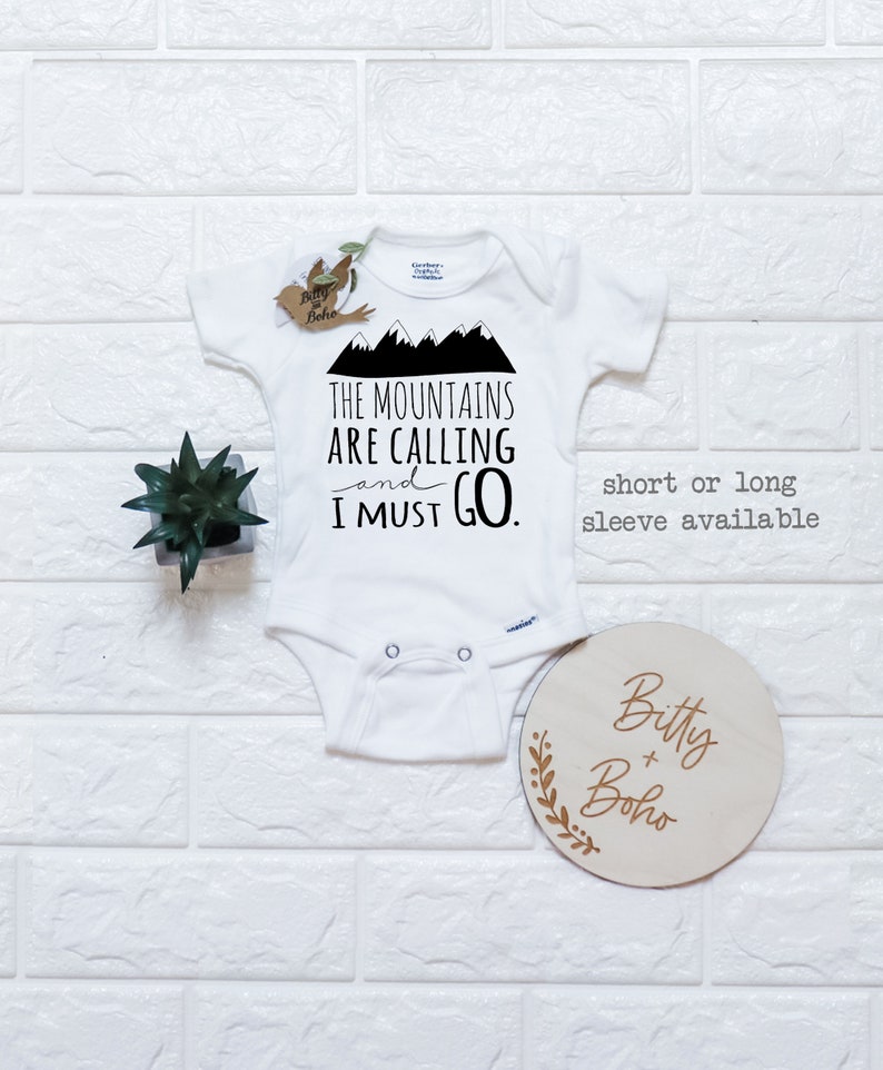 May include: White baby onesie with black text that reads "The Mountains Are Calling and I Must Go."  The onesie has a mountain graphic.  A small wooden sign with the text "Bitty + Boho" is in the lower right corner of the image.