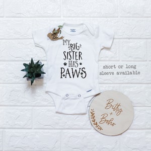 May include: White baby onesie with black text that reads "My Big Sister Has Paws". The onesie is on a white brick background with a small potted plant and a wooden sign that reads "Bitty + Boho".