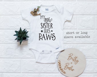 May include: White baby onesie with black text that reads "My Big Sister Has Paws". The onesie is on a white brick background with a small potted plant and a wooden sign that reads "Bitty + Boho".