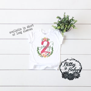K&ouml;nnte beinhalten: Wei&szlig;es T-Shirt mit einer pinkfarbenen Zahl 2 und dem Namen "Emily" in einem Blumenkranz. Das Shirt ist in kurzen oder langen &Auml;rmeln erh&auml;ltlich. Der Text "Bitty and Boho" befindet sich in einem schwarzen Kranz mit Blumen.