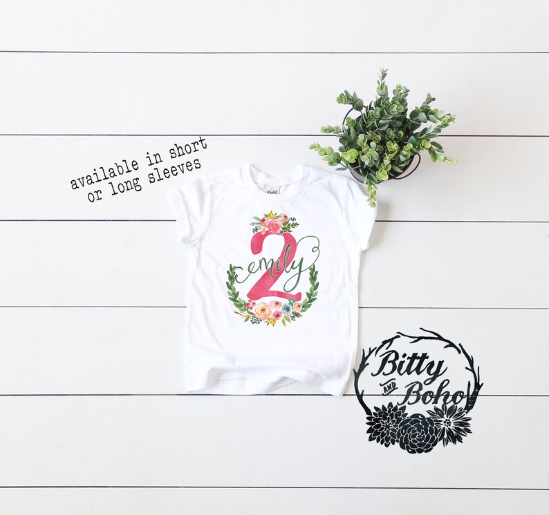 K&ouml;nnte beinhalten: Wei&szlig;es T-Shirt mit einer pinkfarbenen Zahl 2 und dem Namen "Emily" in einem Blumenkranz. Das Shirt ist in kurzen oder langen &Auml;rmeln erh&auml;ltlich. Der Text "Bitty and Boho" befindet sich in einem schwarzen Kranz mit Blumen.