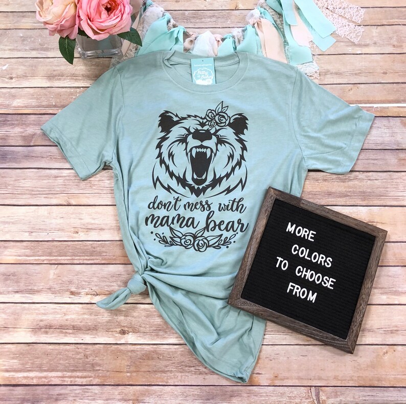 May include: A light blue t-shirt with a black graphic of a bear with the text "don't mess with mama bear". A framed sign next to the shirt reads "more colors to choose from".