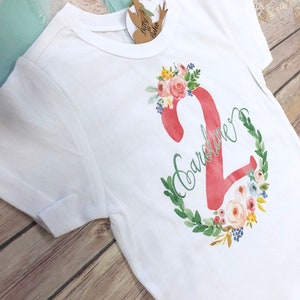 K&ouml;nnte beinhalten: Wei&szlig;es T-Shirt mit einer pinkfarbenen Zahl Zwei, die von einem Blumenkranz umgeben ist. Der Name "Caroline" ist in Kursivschrift auf dem Shirt geschrieben.