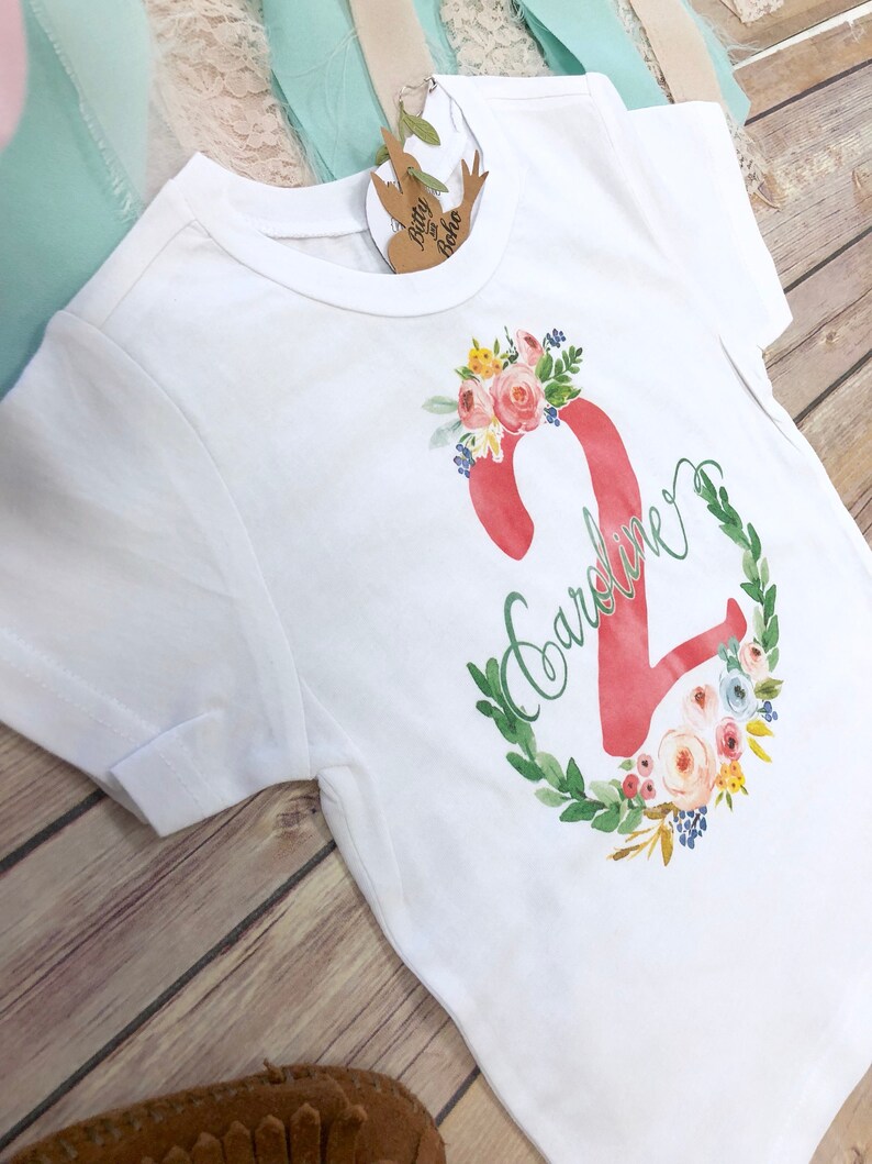 K&ouml;nnte beinhalten: Wei&szlig;es T-Shirt mit einer pinkfarbenen Zahl Zwei, die von einem Blumenkranz umgeben ist. Der Name "Caroline" ist in Kursivschrift auf dem Shirt geschrieben.