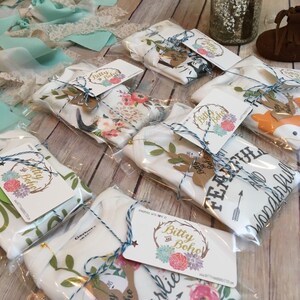 K&ouml;nnte beinhalten: Mehrere Baby-Bodys und -Decken sind in Klarsichtfolie verpackt, mit blauem Garn gebunden und mit Etiketten versehen. Die Etiketten zeigen das "Bitty Boho"-Logo und einen hirschf&ouml;rmigen Ausschnitt. Die Bodys und Decken haben Blumen- und Tiermotive.