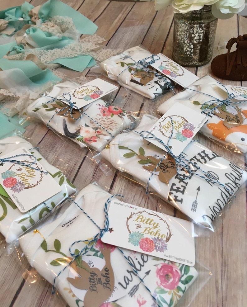K&ouml;nnte beinhalten: Mehrere Baby-Bodys und -Decken sind in Klarsichtfolie verpackt, mit blauem Garn gebunden und mit Etiketten versehen. Die Etiketten zeigen das "Bitty Boho"-Logo und einen hirschf&ouml;rmigen Ausschnitt. Die Bodys und Decken haben Blumen- und Tiermotive.