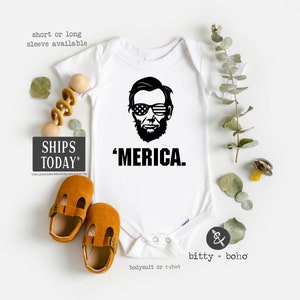 Puede incluir: Un body blanco de manga corta con un gr&aacute;fico negro de Abraham Lincoln usando gafas de sol con la bandera estadounidense y el texto 'MERICA. debajo. La prenda cuenta con un cierre de botones a presi&oacute;n en la parte inferior.