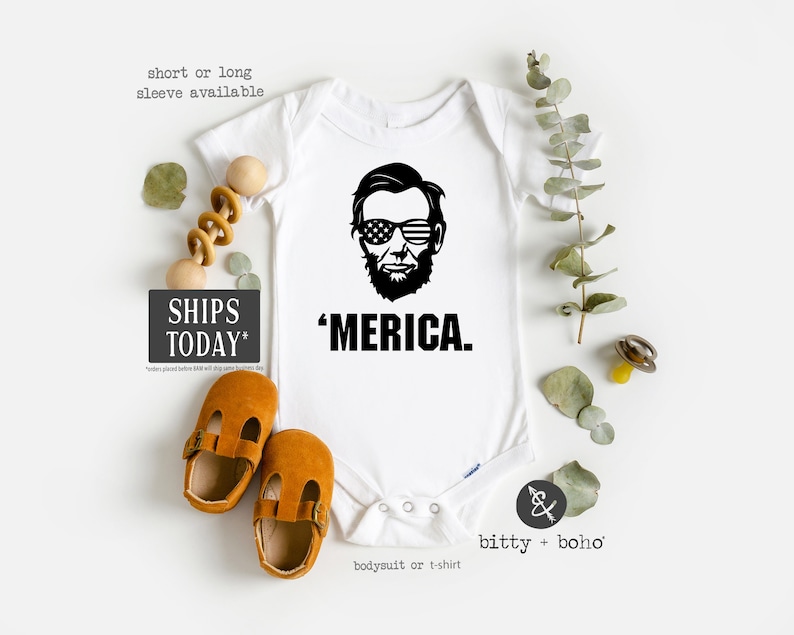 Puede incluir: Un body blanco de manga corta con un gr&aacute;fico negro de Abraham Lincoln usando gafas de sol con la bandera estadounidense y el texto 'MERICA. debajo. La prenda cuenta con un cierre de botones a presi&oacute;n en la parte inferior.