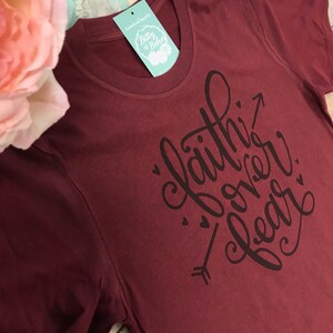 May include: A maroon t-shirt with the phrase "Faith Over Fear" in black script, accompanied by an arrow and small heart accents. The shirt has a crew neck and a tag that reads "Bitty & Boho".