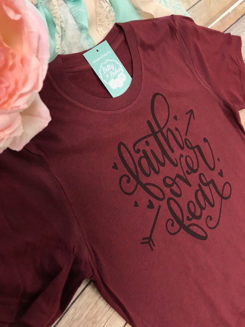 May include: A maroon t-shirt with the phrase "Faith Over Fear" in black script, accompanied by an arrow and small heart accents. The shirt has a crew neck and a tag that reads "Bitty & Boho".