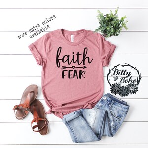 May include: A dusty rose t-shirt with the words "faith" above "fear" in black script, with an arrow and heart design. The shirt is paired with blue jeans and brown sandals. Text on the shirt reads "more shirt colors available".