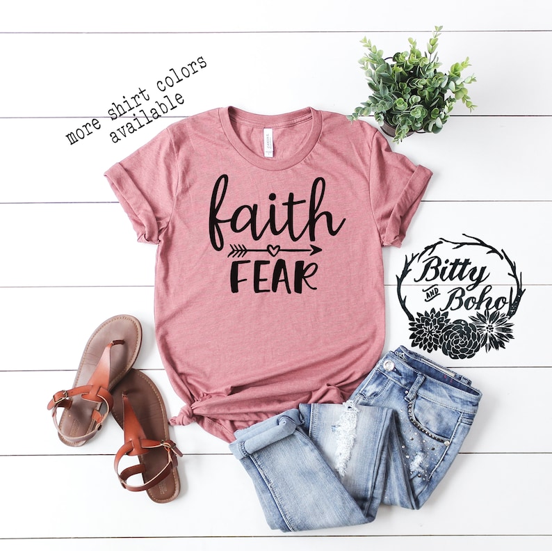 May include: A dusty rose t-shirt with the words "faith" above "fear" in black script, with an arrow and heart design. The shirt is paired with blue jeans and brown sandals. Text on the shirt reads "more shirt colors available".
