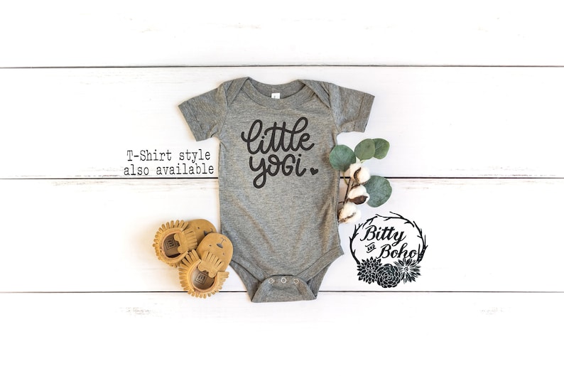 May include: Gray baby onesie with the text "little yogi" in black cursive lettering. The onesie is on a white wooden background with a small cotton plant and eucalyptus leaves.
