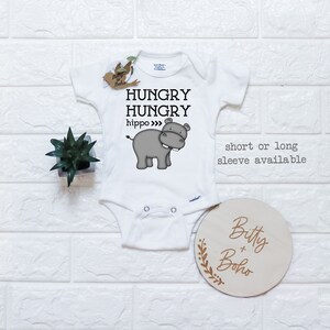 May include: White baby onesie with a gray hippopotamus illustration and the text "Hungry Hungry hippo" printed on the front. The onesie is available in short or long sleeve. A wooden sign with the text "Bitty + Boho" is in the lower right corner of the image.