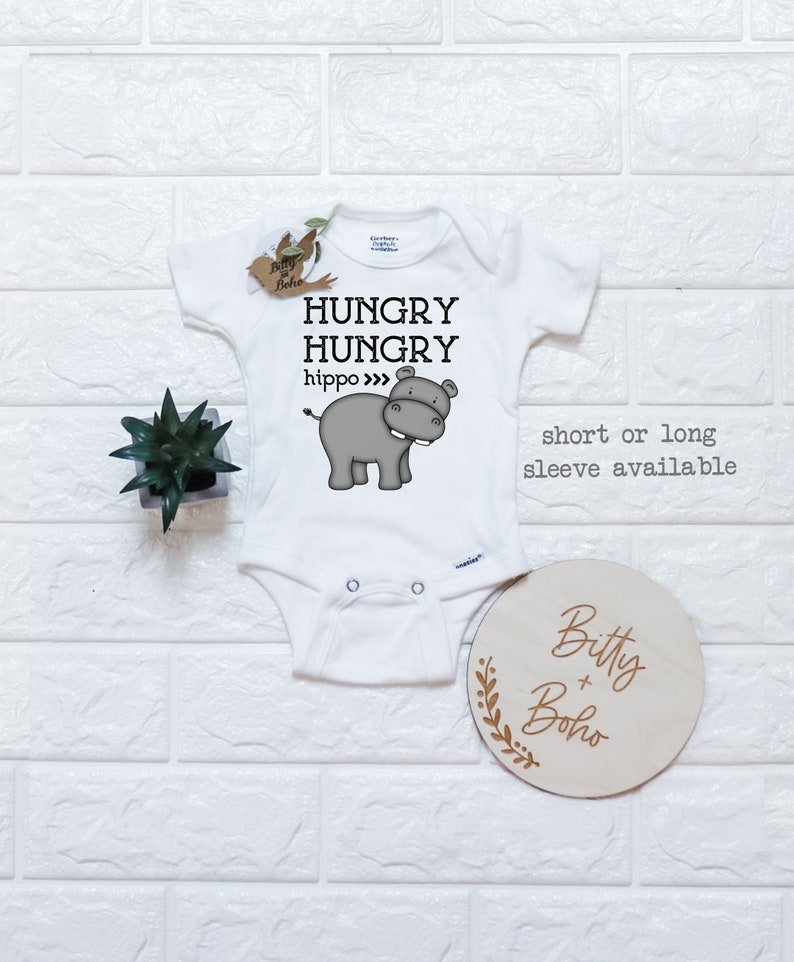 May include: White baby onesie with a gray hippopotamus illustration and the text "Hungry Hungry hippo" printed on the front. The onesie is available in short or long sleeve. A wooden sign with the text "Bitty + Boho" is in the lower right corner of the image.