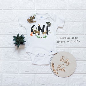 Op de afbeelding: Witte babybody met het woord "ONE" in zwarte letters, versierd met illustraties van bosdieren. Inclusief een houten rond bord met de tekst "Bitty + Boho" en een kleine plant.
