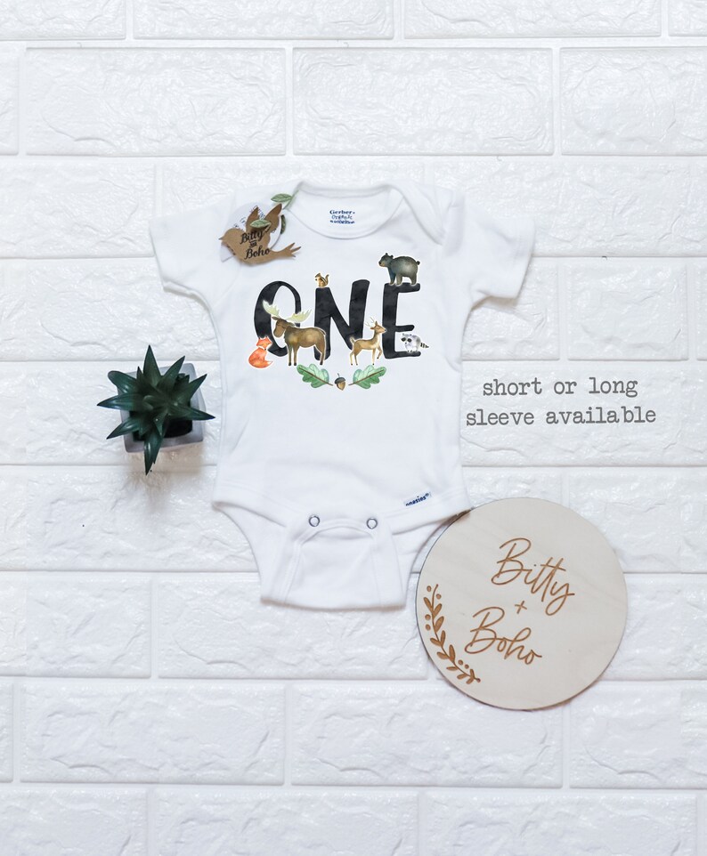 Op de afbeelding: Witte babybody met het woord "ONE" in zwarte letters, versierd met illustraties van bosdieren. Inclusief een houten rond bord met de tekst "Bitty + Boho" en een kleine plant.