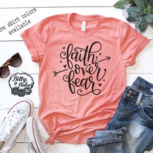 May include: A coral-colored t-shirt with the phrase "faith over fear" in black script, accented with hearts and an arrow. The shirt is displayed with blue jeans, white sneakers, and sunglasses.