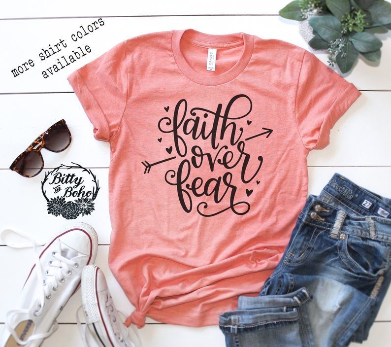 May include: A coral-colored t-shirt with the phrase "faith over fear" in black script, accented with hearts and an arrow. The shirt is displayed with blue jeans, white sneakers, and sunglasses.