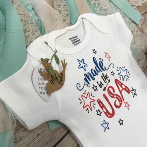 Puede incluir: Body de beb&eacute; blanco con el texto "Made in the USA" en rojo y azul, decorado con estrellas y una flecha. El body tiene mangas cortas y una etiqueta de "Bitt and Boho". La marca Gerber es visible en el cuello.