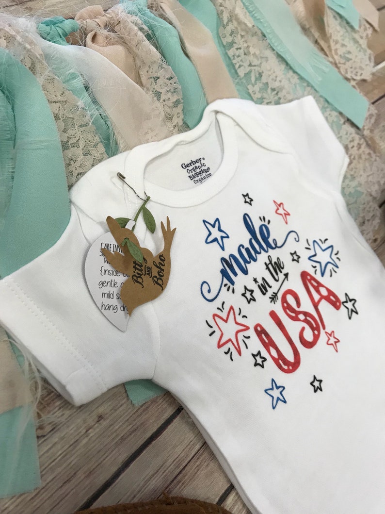 Puede incluir: Body de beb&eacute; blanco con el texto "Made in the USA" en rojo y azul, decorado con estrellas y una flecha. El body tiene mangas cortas y una etiqueta de "Bitt and Boho". La marca Gerber es visible en el cuello.