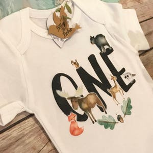 Op de afbeelding: Witte babybody met een bosdierontwerp. Het woord "ONE" is gedrukt in grote zwarte letters, met een vos, eland, hert en andere dieren. Een label met de woorden "Bitty & Boho" is bevestigd.