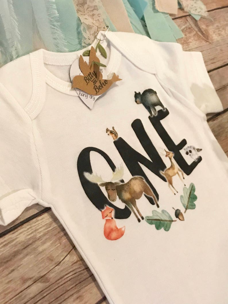 Op de afbeelding: Witte babybody met een bosdierontwerp. Het woord "ONE" is gedrukt in grote zwarte letters, met een vos, eland, hert en andere dieren. Een label met de woorden "Bitty & Boho" is bevestigd.