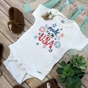 Puede incluir: Un body blanco para beb&eacute; con el texto "Made in the USA" en rojo y azul, decorado con estrellas. Mocasines de beb&eacute; de ante marr&oacute;n, un par de calcetines marrones con tem&aacute;tica de zorro y una peque&ntilde;a planta suculenta tambi&eacute;n son visibles.