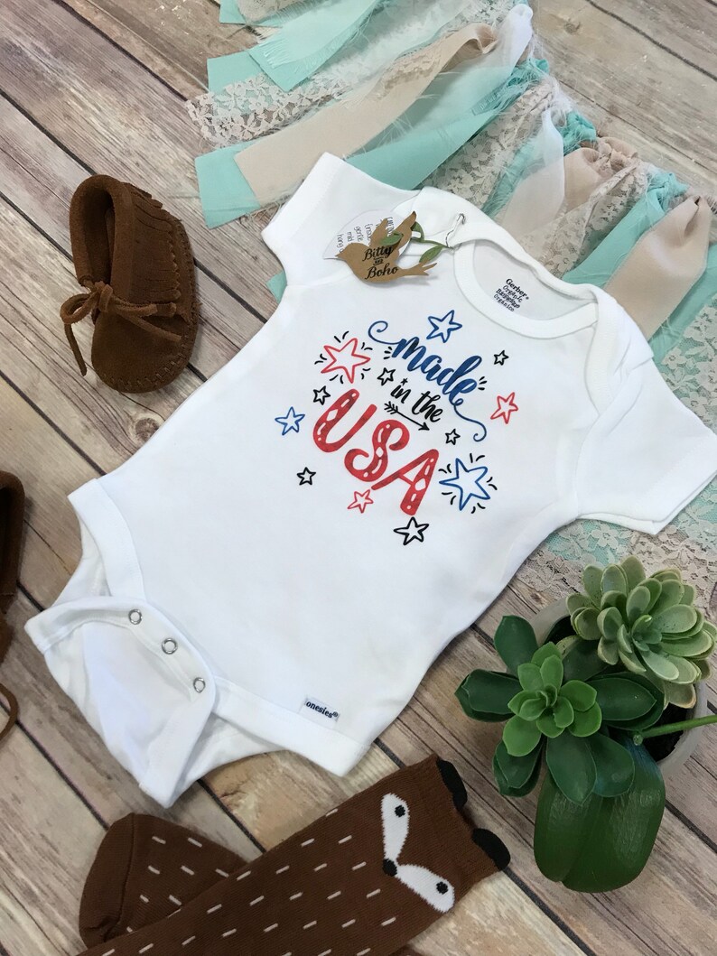 Puede incluir: Un body blanco para beb&eacute; con el texto "Made in the USA" en rojo y azul, decorado con estrellas. Mocasines de beb&eacute; de ante marr&oacute;n, un par de calcetines marrones con tem&aacute;tica de zorro y una peque&ntilde;a planta suculenta tambi&eacute;n son visibles.