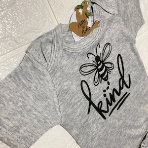 May include: A gray baby onesie with a black graphic of a bee and the word "kind". The onesie has short sleeves and snap closures. A tag with the words "Bitty and Boho" is attached to the onesie.