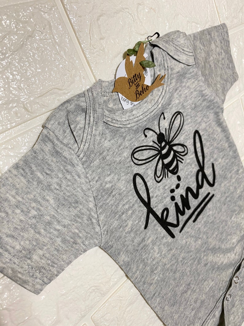 May include: A gray baby onesie with a black graphic of a bee and the word "kind". The onesie has short sleeves and snap closures. A tag with the words "Bitty and Boho" is attached to the onesie.