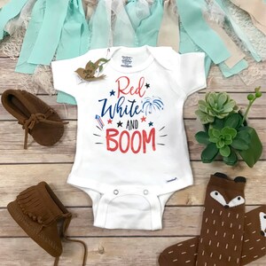 May include: A white short-sleeved baby bodysuit featuring the red and blue text "Red White AND BOOM" with graphic illustrations of a firework and stars. The garment includes a small tag, two metal snaps at the crotch, and a printed neck label.