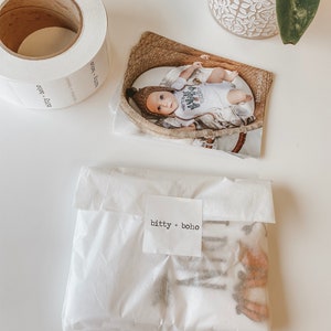 以下が含まれることがあります： 「bitty + boho」の文字が入った白いラベルのロールと、「bitty + boho」と書かれたステッカーが付いた包装されたパッケージ。 籐のバスケットに入った赤ちゃんの写真も写っています。
