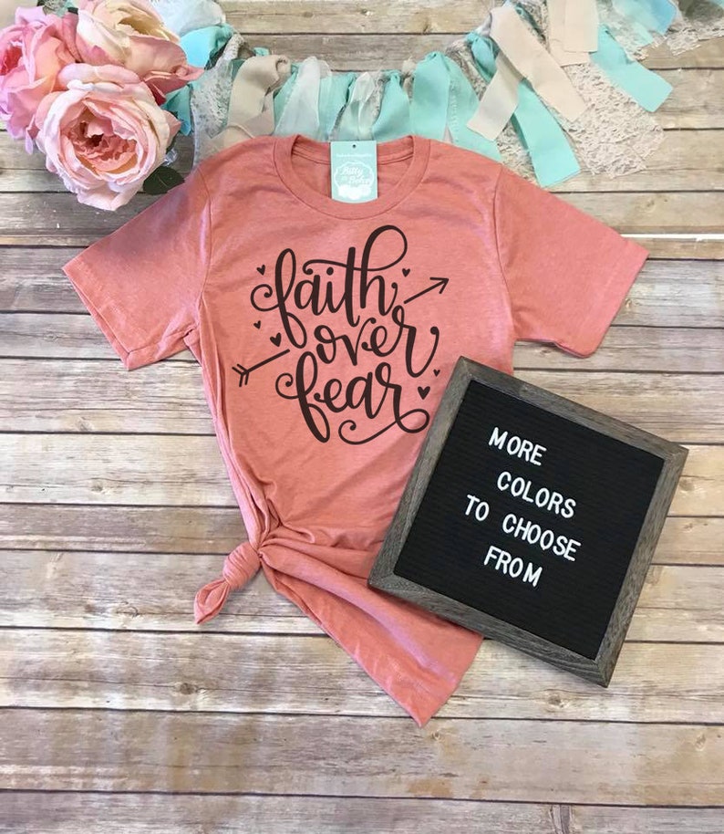 May include: A coral-colored t-shirt with the words "faith over fear" in black script, accented with hearts and an arrow. A small sign reads "MORE COLORS TO CHOOSE FROM". The shirt is knotted at the waist.