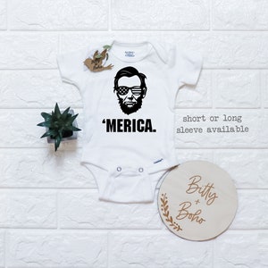 Puede incluir: Un body blanco para beb&eacute; con un gr&aacute;fico negro de Abraham Lincoln usando gafas de sol con el patr&oacute;n de la bandera estadounidense. Debajo del gr&aacute;fico est&aacute; el texto 'MERICA. La prenda tiene mangas cortas y broches met&aacute;licos en la entrepierna.
