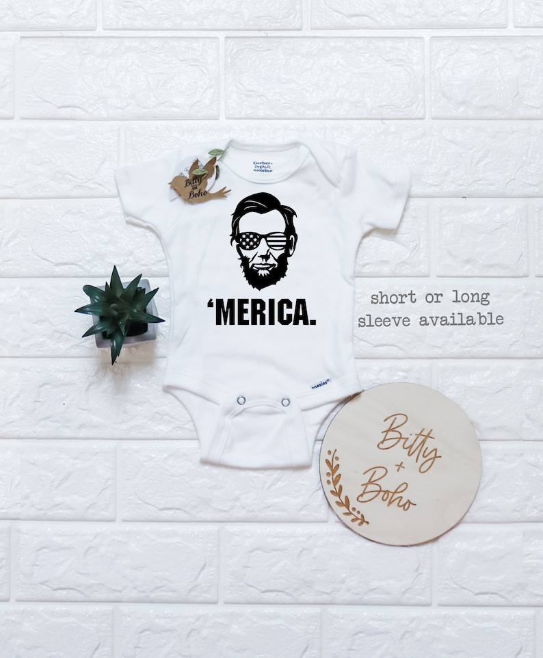 Puede incluir: Un body blanco para beb&eacute; con un gr&aacute;fico negro de Abraham Lincoln usando gafas de sol con el patr&oacute;n de la bandera estadounidense. Debajo del gr&aacute;fico est&aacute; el texto 'MERICA. La prenda tiene mangas cortas y broches met&aacute;licos en la entrepierna.