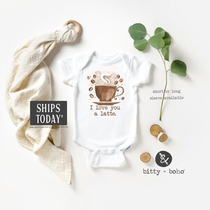 May include: White baby onesie with a brown coffee cup illustration and the text "I love you a latte."  The onesie is surrounded by a white background with a wooden ring, a beige muslin swaddle, and a sprig of greenery.