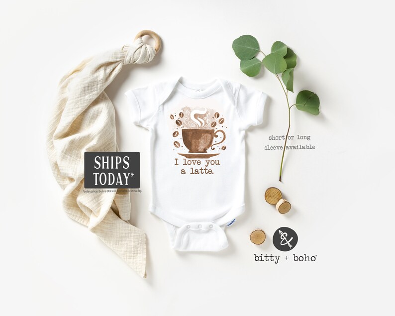 May include: White baby onesie with a brown coffee cup illustration and the text "I love you a latte."  The onesie is surrounded by a white background with a wooden ring, a beige muslin swaddle, and a sprig of greenery.
