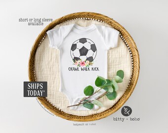 May include: White baby bodysuit with a soccer ball graphic and floral accents. Text reads "CRAWL. WALK. KICK." The bodysuit is displayed in a woven basket with greenery. Short or long sleeve options are available.